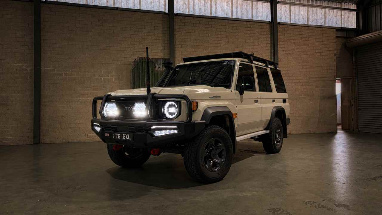 A beige off-road SUV with black trim, rugged tires, roof rack, and front bull bar parked inside an industrial warehouse. The vehicle is photographed from a low front three-quarter angle, with headlights glowing against a darker, moody atmosphere and soft overhead lighting reflecting on the polished concrete floor.